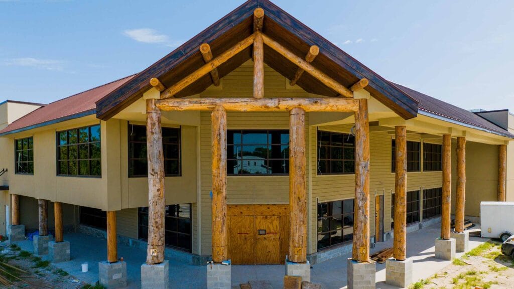 A two-story building under construction featuring large wooden log columns, unfinished entryway, and extensive glass windows under a sloped roof.