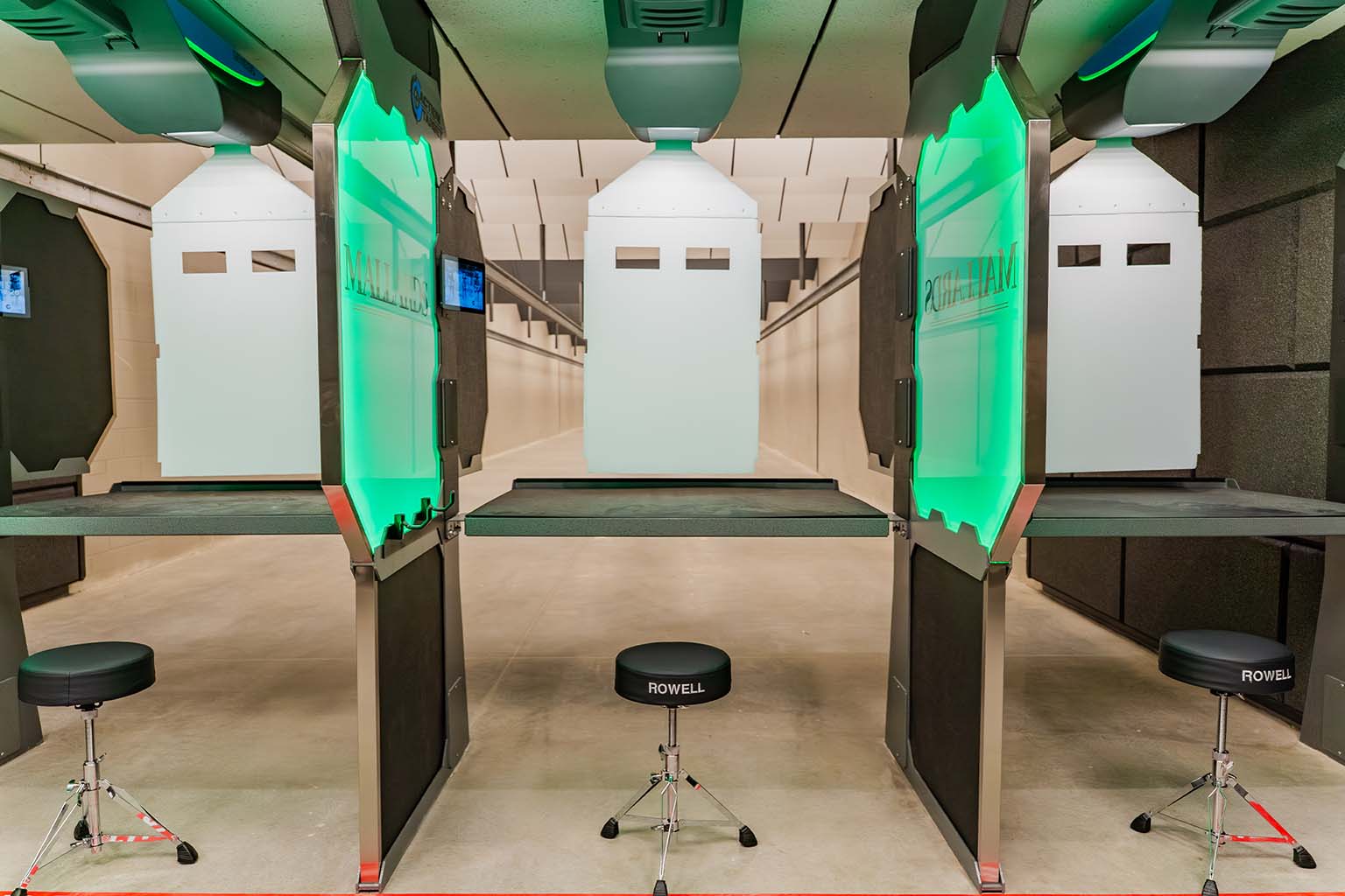 Modern indoor shooting range featuring three booths, comfortable stools, and target boards set up in the distance.