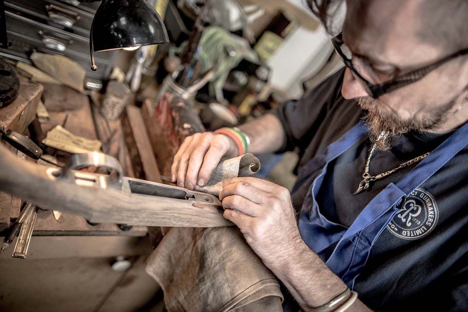 A craftsman in glasses and a blue apron works on a wooden project at a workbench for the John Rigby Company.
