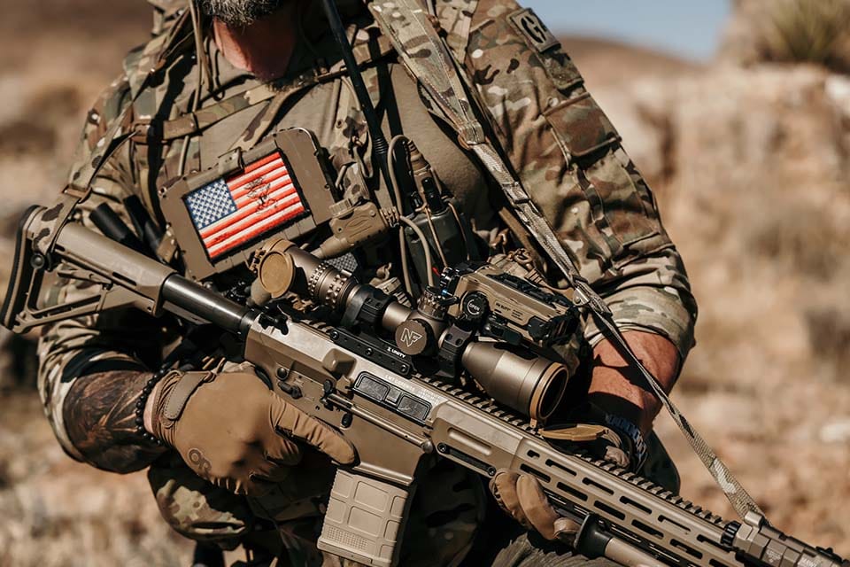 A person in camouflage military gear holding an LMT Defense rifle, with tactical equipment and an American flag patch attached to their vest outdoors.